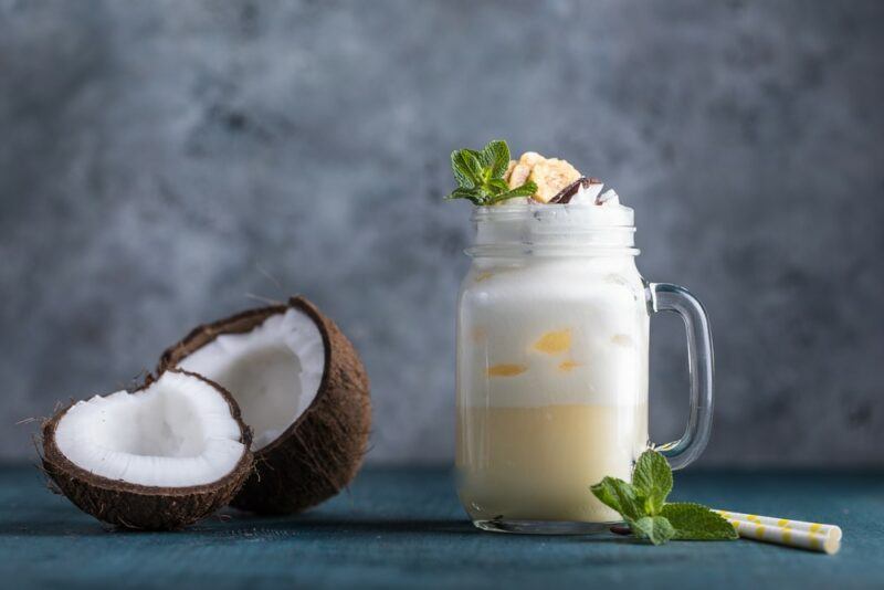 A mason jar with a handle containing a batida cocktail topped with cream or froth, next to two coconut halves