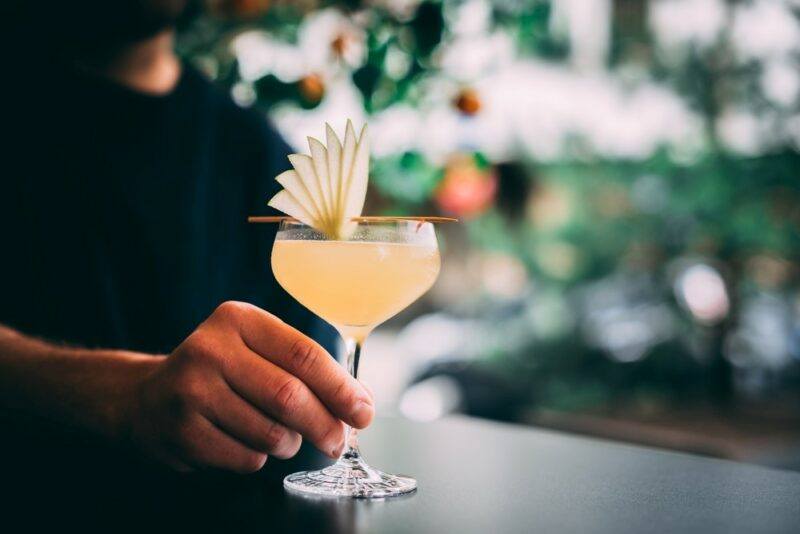 A bartender with a cocktail called For Sake's Sake that has been garnished with a fan of apple slices. There's an out of focus bar in the background