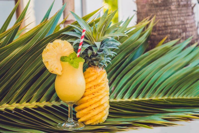 A tropical cocktail called My Thai, garnished with a large piece of pineapple, next to a pineapple, in front of large pineapple fronds