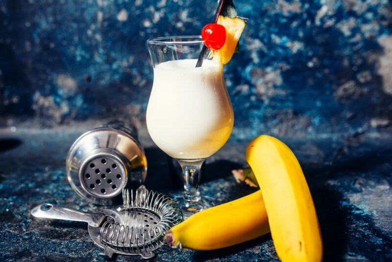 A hurricane glass with a bananalada cocktail next to some bananas, a strainer, and a cocktail shaker