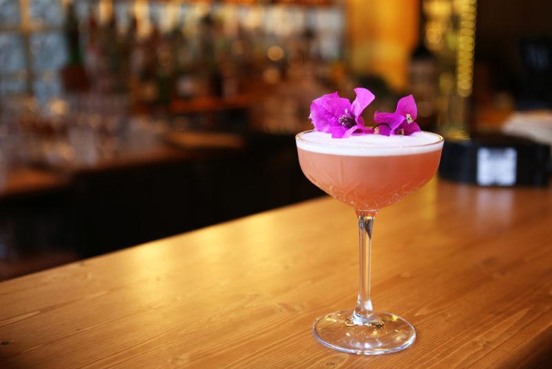 A cocktail in a glass on a bar with a purple flower as a garnish