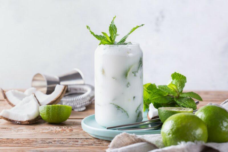 A tall coconut gin fizz cocktail surrounded by limes, coconut, and some bar equipment