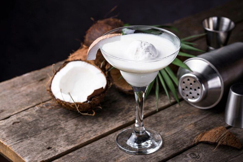 A wooden table with a coconut margarita, next to a cocktail shaker, a leaf, and coconuts