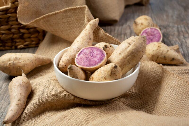 A white bowl containing Okinawan purple sweet potatoes, with a few more sweet potatoes scattered alongside