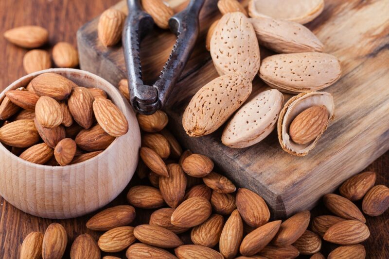 A wooden board with unshelled almonds, with more almonds scattered on the table and in a small bowl