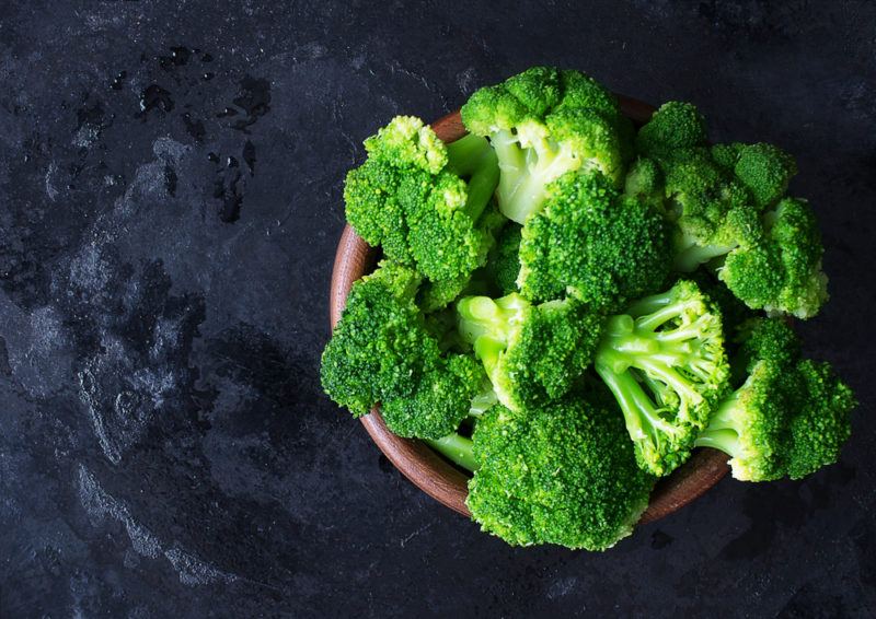 A wooden bowl containing bright green broccoli on a blue table