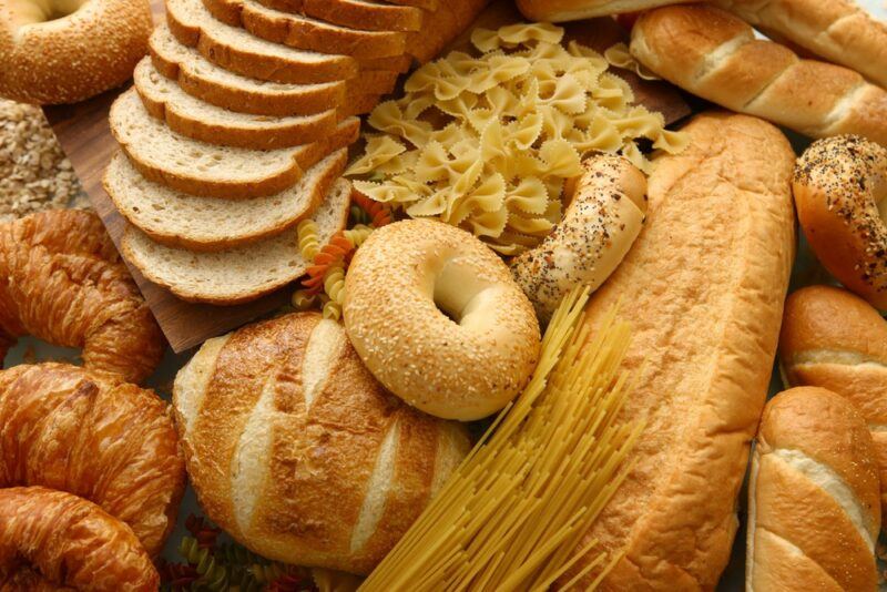 A large collection of gluten containing foods, including loaves of bread, pasta, sliced bread, bagels, and croissants.