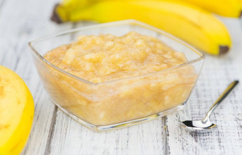 A hite wooden table with a glass jar containing mashed banana, next to a spoon and some fresh bananas