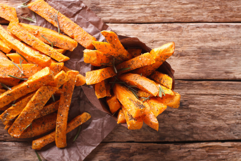 A wooden table or deck with a collection of sweet potato fries, some standing upright in a container and others lying flat