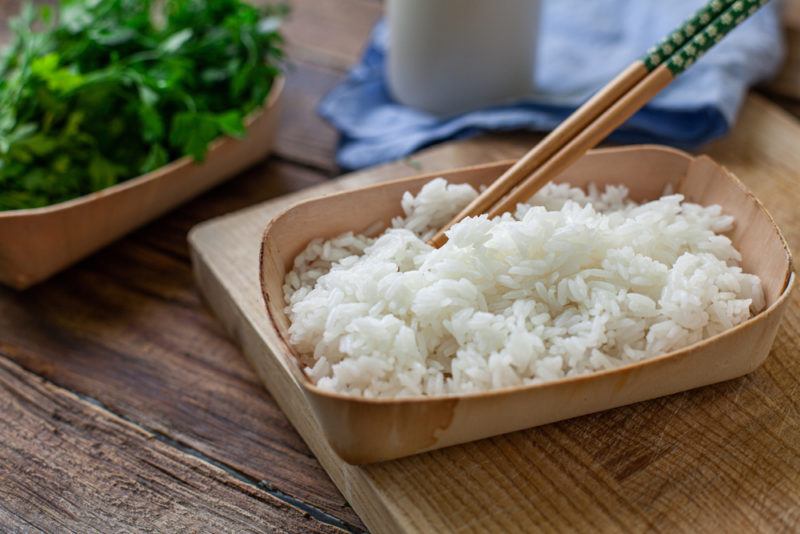 A small brown container with steamed rice and chopsticks on a wooden board, with a container of greens in the background