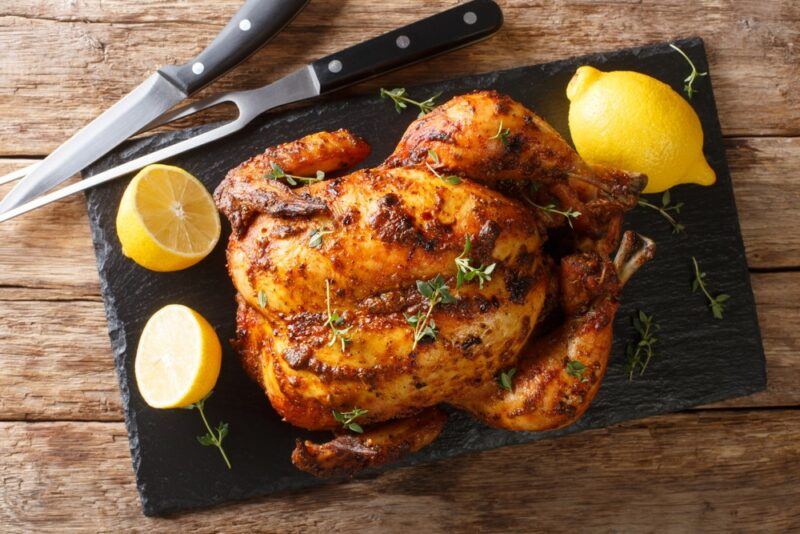 A black slate with a whole cooked rotisserie chicken, with lemons and carving tools