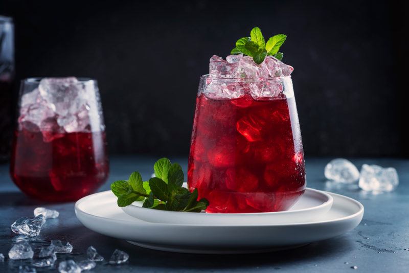 Two glasses of a cranberry tequila punch. One is on a white plate and there is ice scattered around