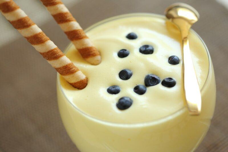 A glass containing a creamy treat called kogel mogel, with two straws, a spoon, and some blueberries