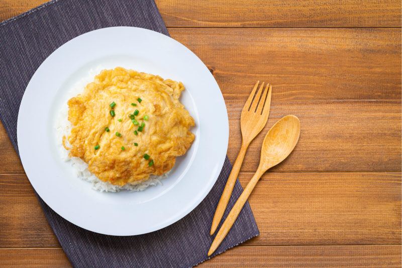 A fried Thai omelet on rice and a white plate, next to a fork and a spoon