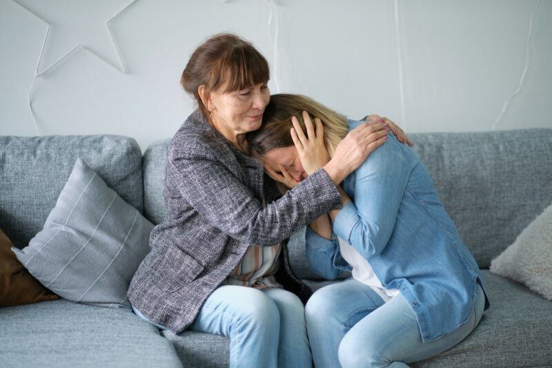 A crying woman sitting on a couch with her mother and being comforted. There's a star on the wall in the background and gray cushions on the couch