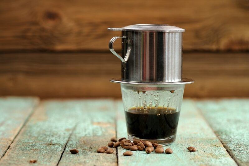 A cup of coffee in Vietnam with the filter on top and coffee beans on the table