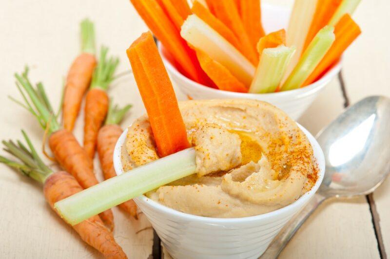 Two white cups, one filled with veggie sticks and the other with hummus, which has a couple of veggie sticks as well. There are fresh baby carrots on the table as well, along with a spoon