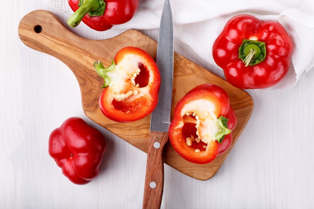 A cutting board with a red pepper that's been sliced in half and a knife, with a few red peppers around the outside