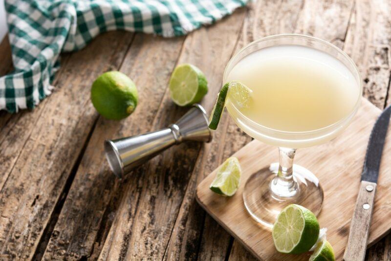 A single daiquiri cocktail on a wooden board, surrounded by limes, next to a cocktail jigger