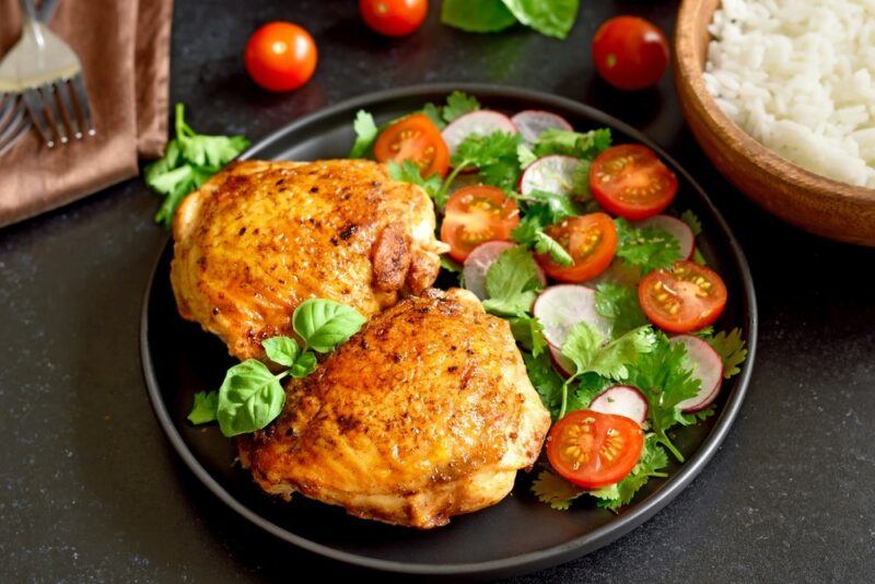 A dark plate with chicken thighs and a tomato and radish based salad