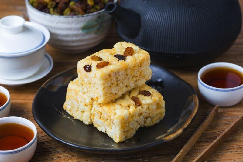 A dark plate containing three pieces of Chinese sachima with sultanas on top, next to various bowls, some of which contain a sweet syrup