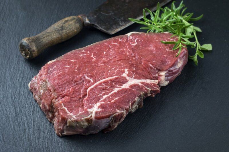 A black board or table with a raw piece of bottom round steak, rosemary, and a cooking tool