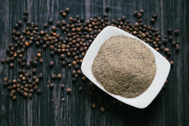 A dark wooden table with peppercorns scattered on it, along with a white dish with freshly ground pepper