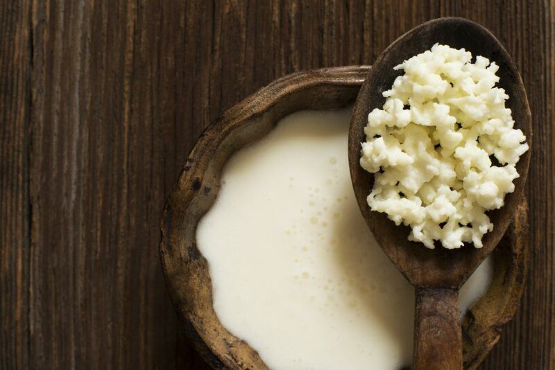 A dark wooden table with a wooden bowl of kefir and a wooden spoon filled with the grains