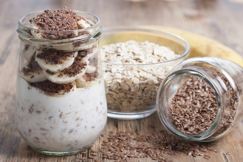 A glass jar containing a a flax seed dessert with cocoa and bananas, next to flax seeds, other seeds, and a banana