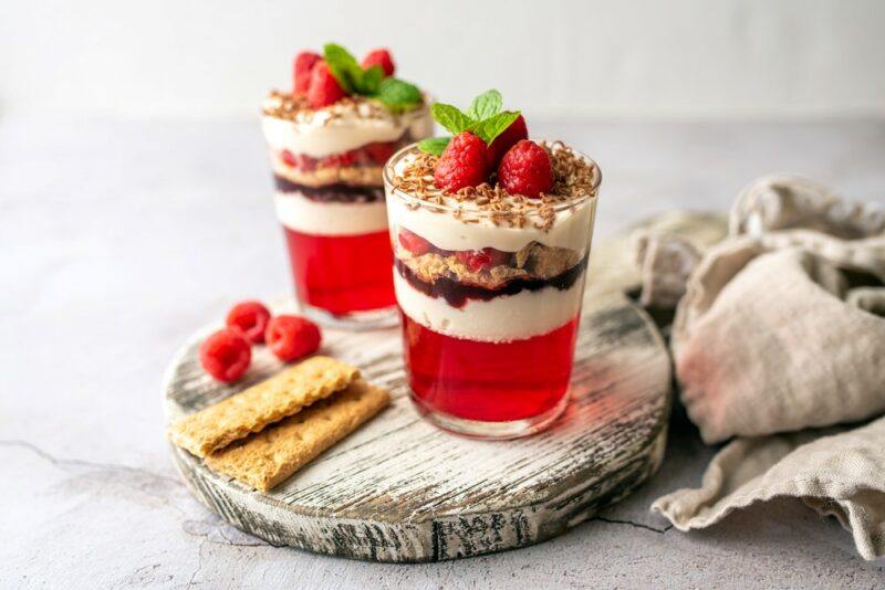 Two glasses containing desserts made using layers of red jello, cream, chocolate, and more