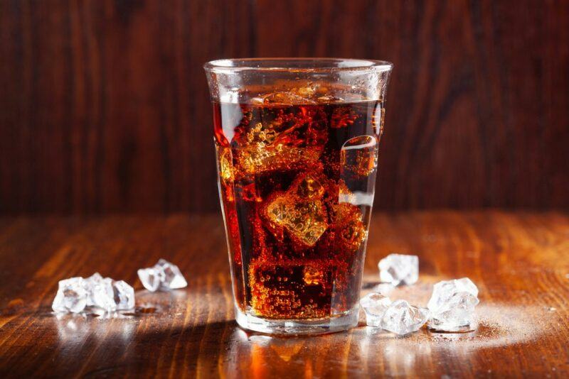 A glass containing a dirty Doctor Pepper cocktail next to ice on a wooden table