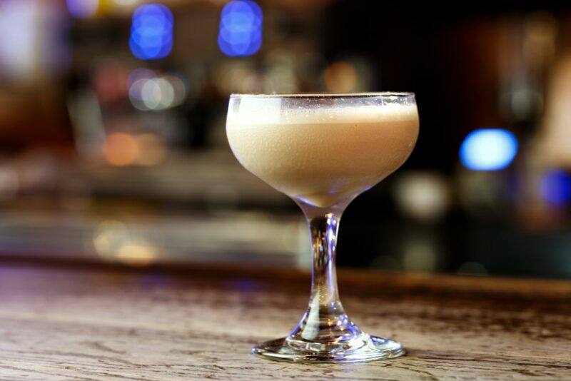 A coupe glass containing a dirty white mother cocktail against an out of focus bar