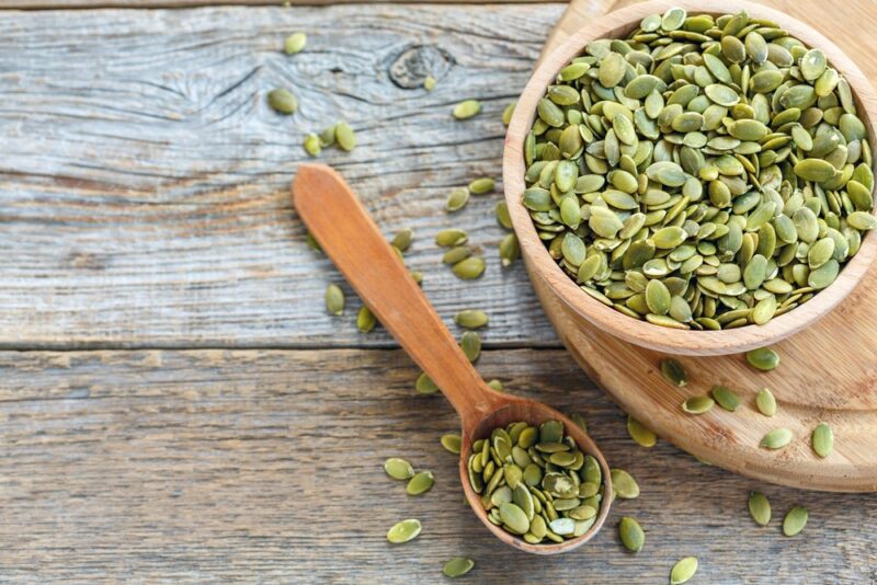 A wooden bowl filled with pumpkin seeds, next to a wooden spoon with more of the seeds