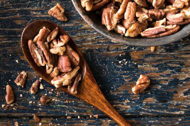 A bowl of pecans next to a brown spoon with more pecans