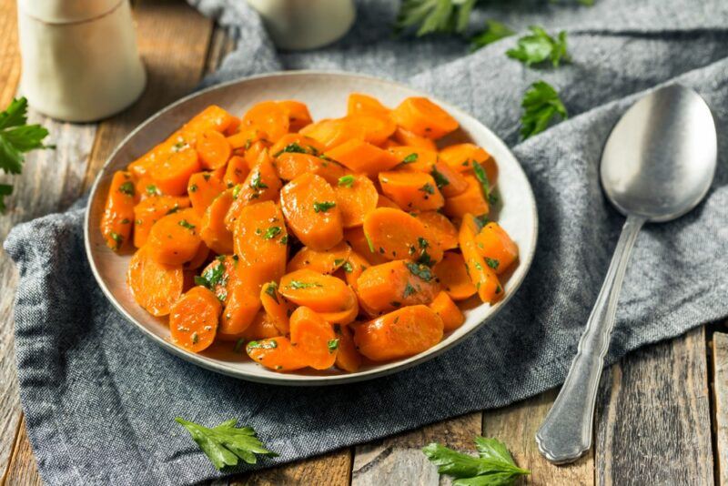 A plate with sliced pieces of cooked carrot on a gray cloth next to a spoon