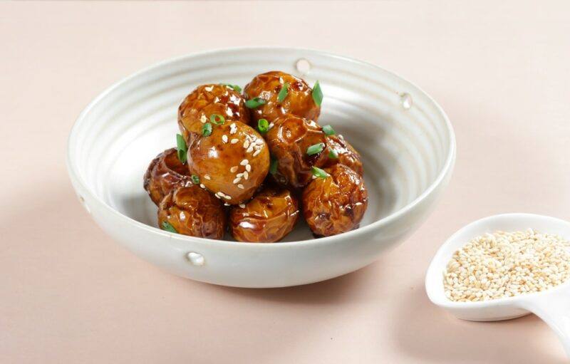 A light colored background with a white dish that contains Korean braised potatoes. These have been made using soy sauce and there is a small dish of sesame seeds next to the bowl.