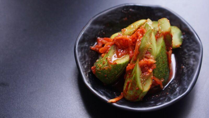 A dark colored table with a black dish containing Korean kimchi stuffed cucumbers.