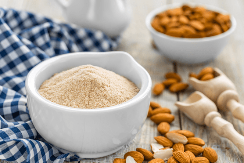 A white bowl containing almond flour, plus almonds on the table and in a small white bowl. There is also a blue and white checkered cloth and two wooden scoops.