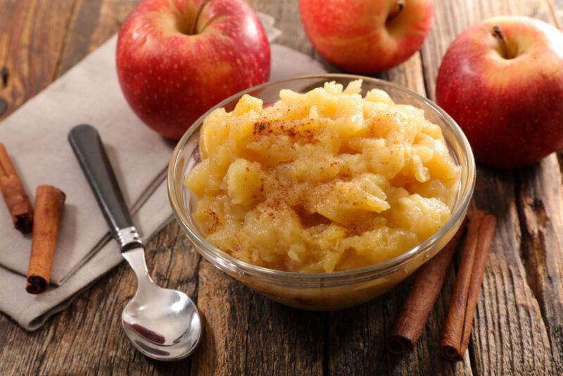 A glass bowl of apple sauce surrounded by red apples, next to a spoon and some cinnamon sticks