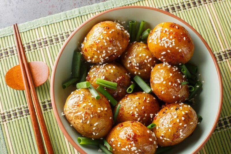 A large bowl containing braised potatoes with sesame seeds, next to chopsticks on a mat