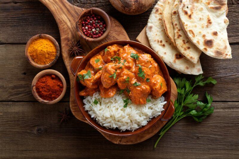 A brown dish with butter chicken and rice, with small bowls of spices and some naan bread around the outside