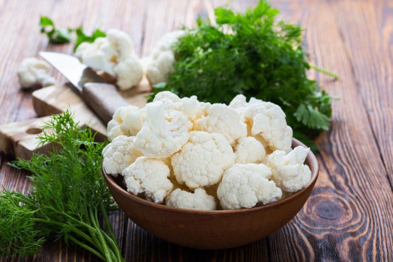 A brown dish of cauliflower on a wooden table, next to some leafy greens.