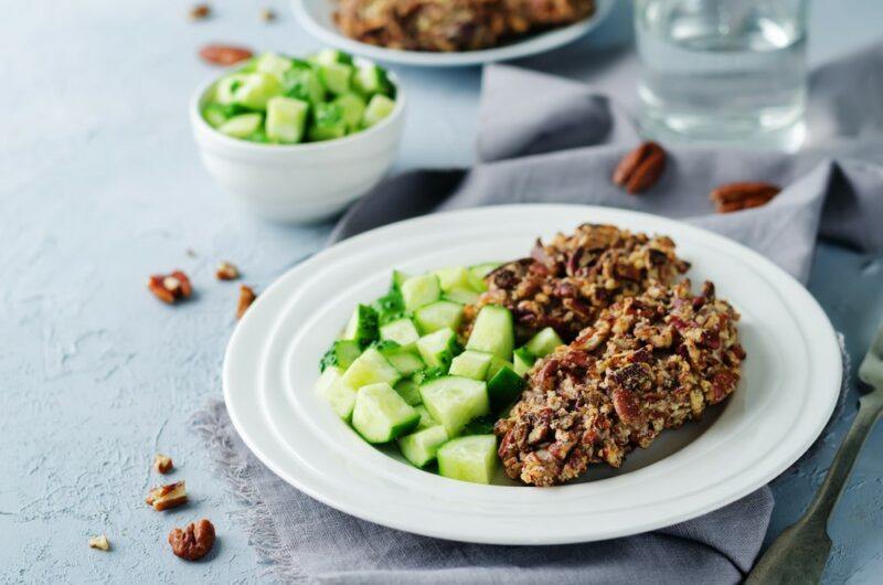 A white plate with a pecan encrusted chicken, next to chopped cucumber