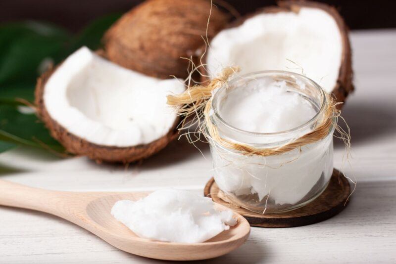 A jar of coconut oil, next to a wooden spoon of coconut oil and some coconut halves