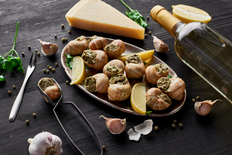 An oval black plate with cooked escargot and lemon, next to a block of cheese and a bottle of white wine