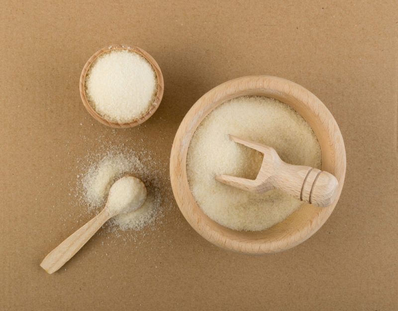 A small brown bowl of gelatin powder with a scoop, along with a smaller container of the powder and a wooden spoon with the powder too