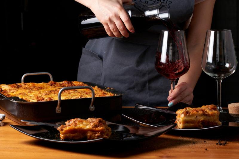 A dish of lasagna next to a chef pouring two glasses of red wine