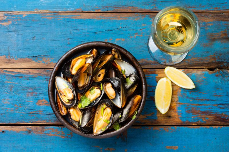 A black dish of mussels on a blue table next to lemon wedges and a glass of wine