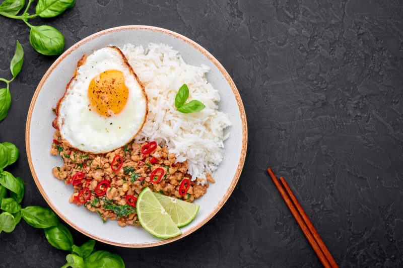 A white plate with pad krapow, rice, limes, and a fried egg