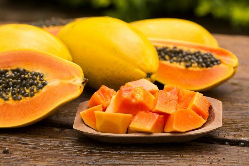 A small dish of papaya pieces next to a few whole papayas and some that have been cut in half